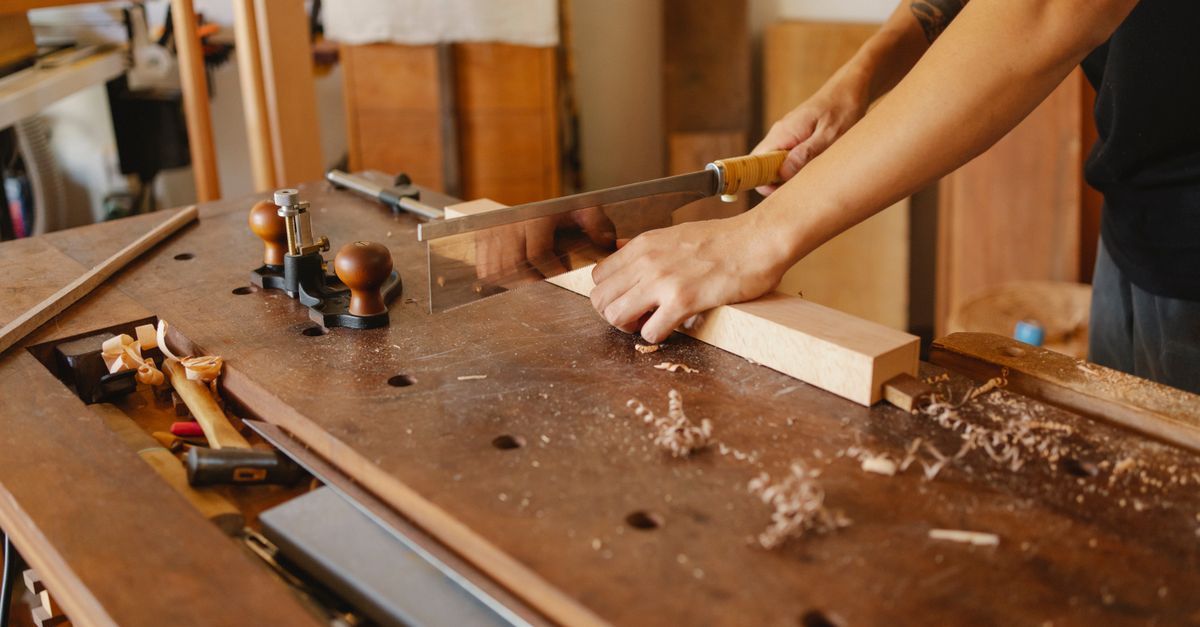 Crop anonymous craftsman with tattoo sawing wooden board with hacksaw at workplace in carpentry workshop